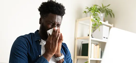 A man blowing his nose while working on a laptop at his desk at home.