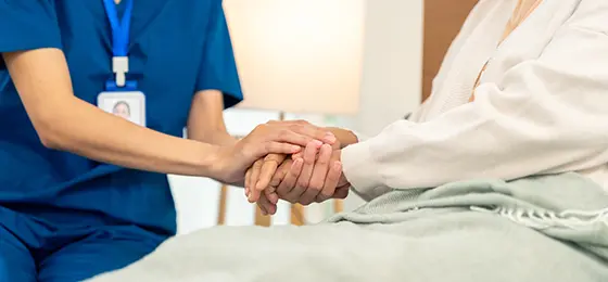 Woman caregiver holding hands with senior woman who is sitting in bed.