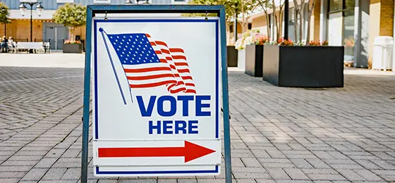 Close-up of "Vote Here" directional sign with US flag at polling station.