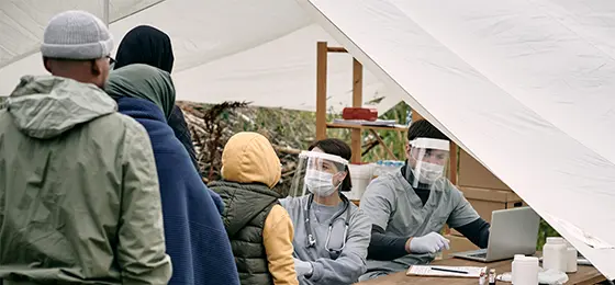 Medical specialists in protective masks sitting at table in tent and checking health of migrants standing in line.