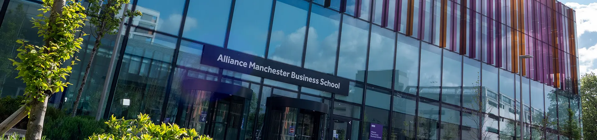 Exterior view of Alliance Manchester Business School building with glass façade and colorful vertical panels, surrounded by greenery on a sunny day.
