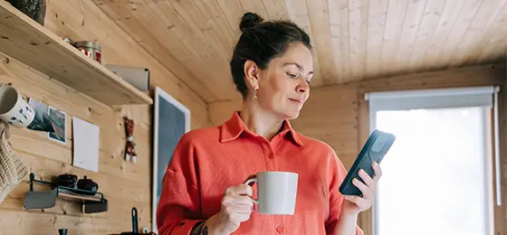 A woman looking at her phone and holding a cup of tea in a kitchen with wooden panels.