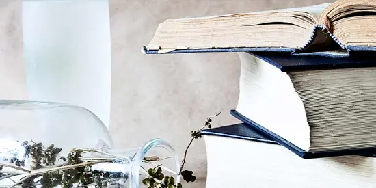 Open books stacked beside a glass bottle with dried flowers spilling out on a light surface