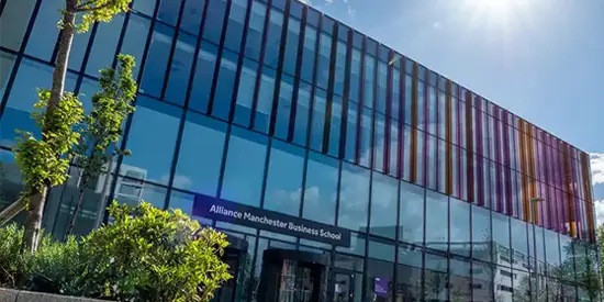 Exterior view of Alliance Manchester Business School building with glass fa&ccedil;ade and colorful vertical panels, surrounded by greenery on a sunny day.