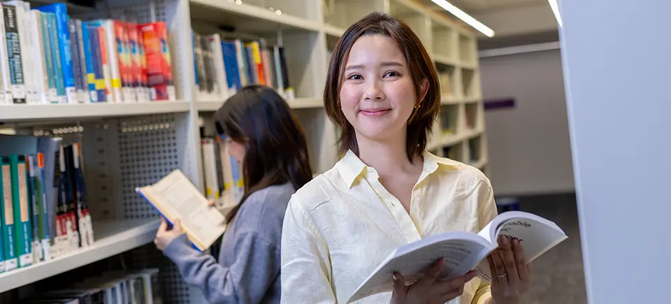 A female student standing between library bookshelves holding an open book.