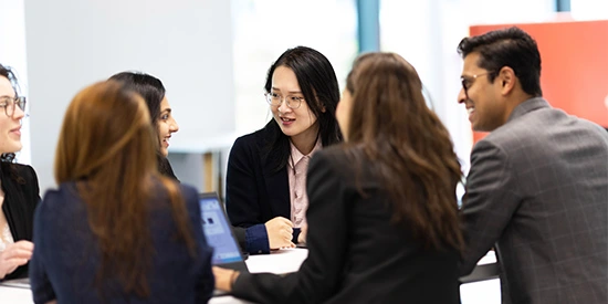 A group of MBA students sitting around a table, engaged in a discussion, with laptops and notebooks in front of them