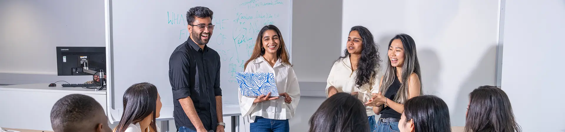 Four students presenting to their classmates in front of two large whiteboards.