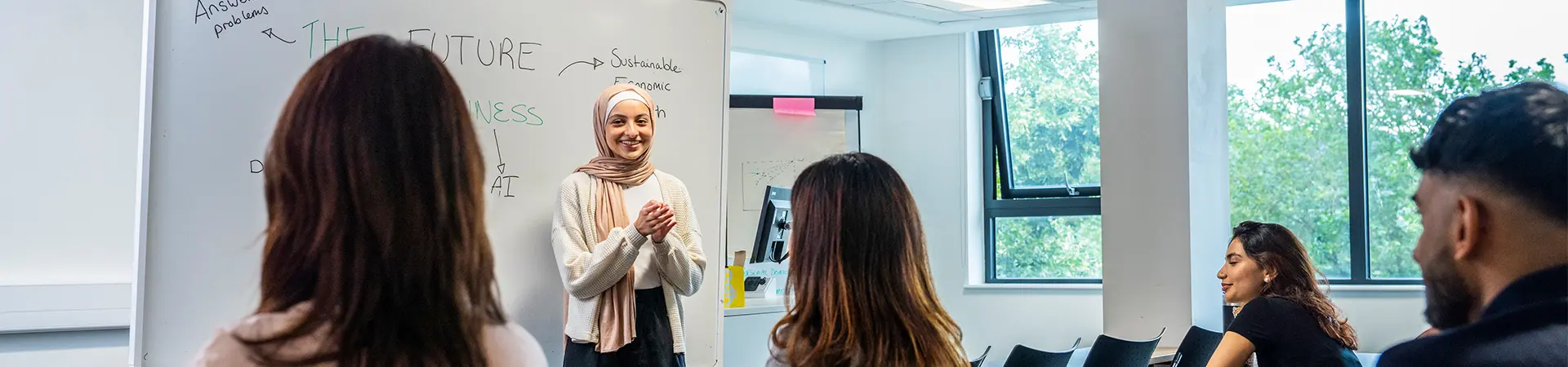 A female student presenting to four classmates in front of a large whiteboard.