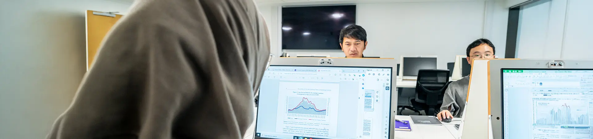 A student working on data on a laptop with two other students in the background.