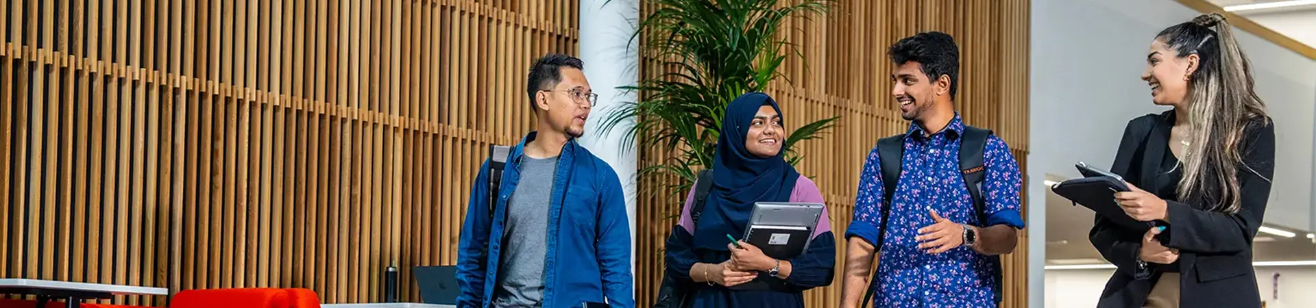 Four students walking through a modern hallway at Alliance Manchester Business School, smiling and talking while carrying laptops and backpacks.