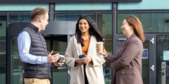 Three students outside the AMBS building chatting and drinking coffee