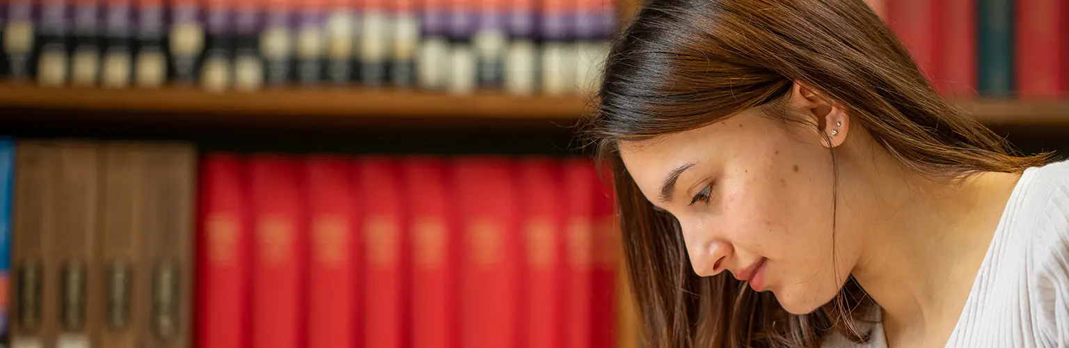 Student concentrating in a library, with shelves of red and multicolored books in the background.
