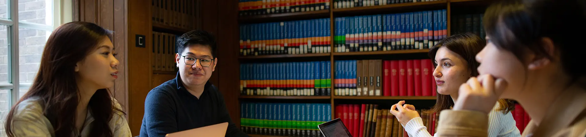 Four students working together in a traditional library setting with books in the background.