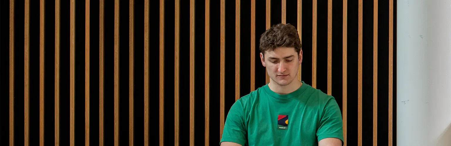 Student in a green shirt sitting indoors against a modern wooden panel wall, focused on studying.