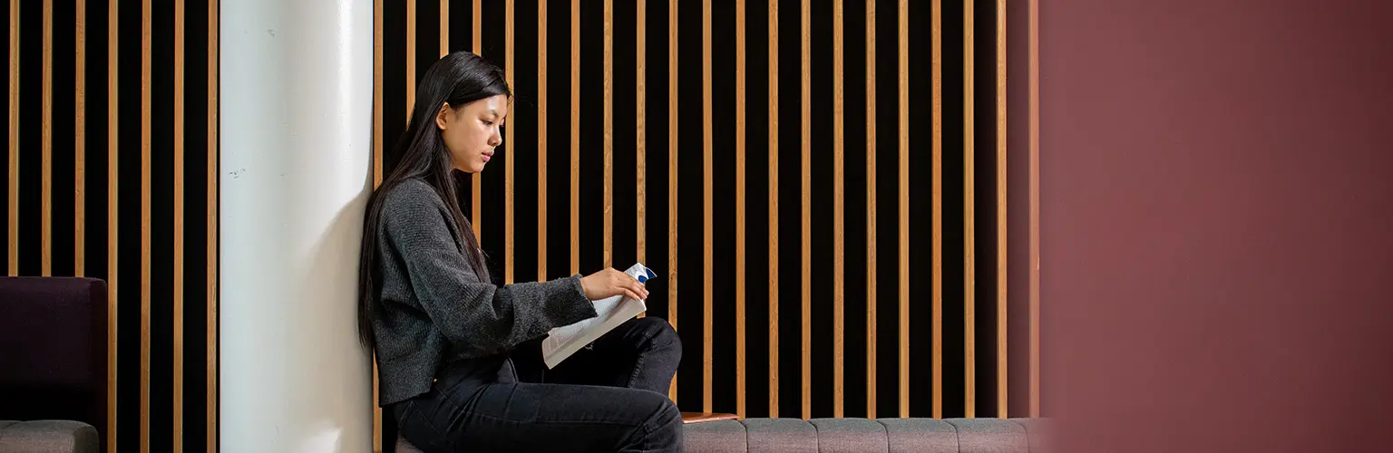 Student sitting indoors against a modern wooden panel wall, reading a book with focused attention.