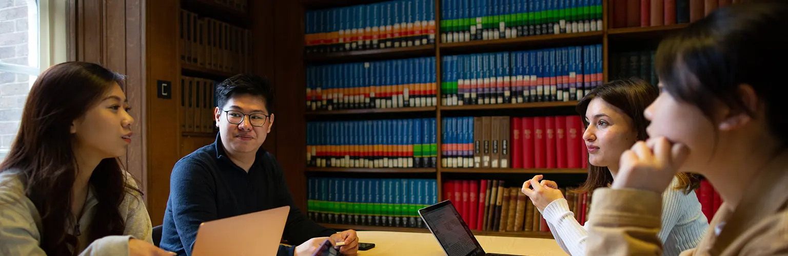 Four students sitting around a table in a library, engaged in discussion with laptops and bookshelves in the background.