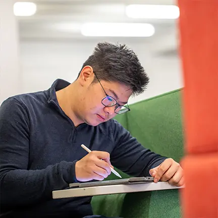 Student using a stylus to work on a tablet while seated in a modern study area with green and red cushioned furniture.