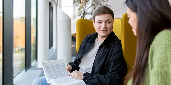 A male student holding an open book while talking to a female student who is sitting beside him in the AMBS library.