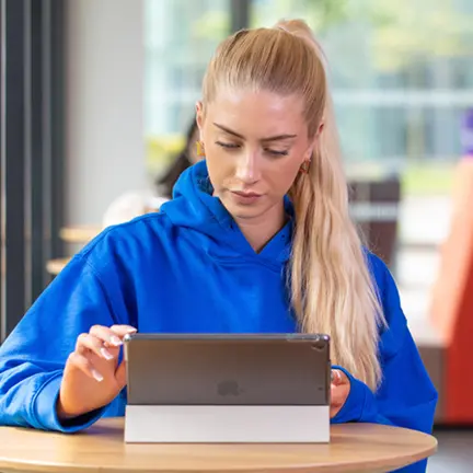 Woman in a blue hoodie using a tablet at a round table in a modern indoor setting.