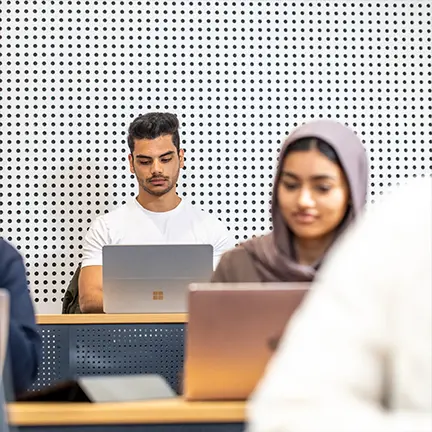 Students working on laptops in a modern classroom with a perforated white wall background.