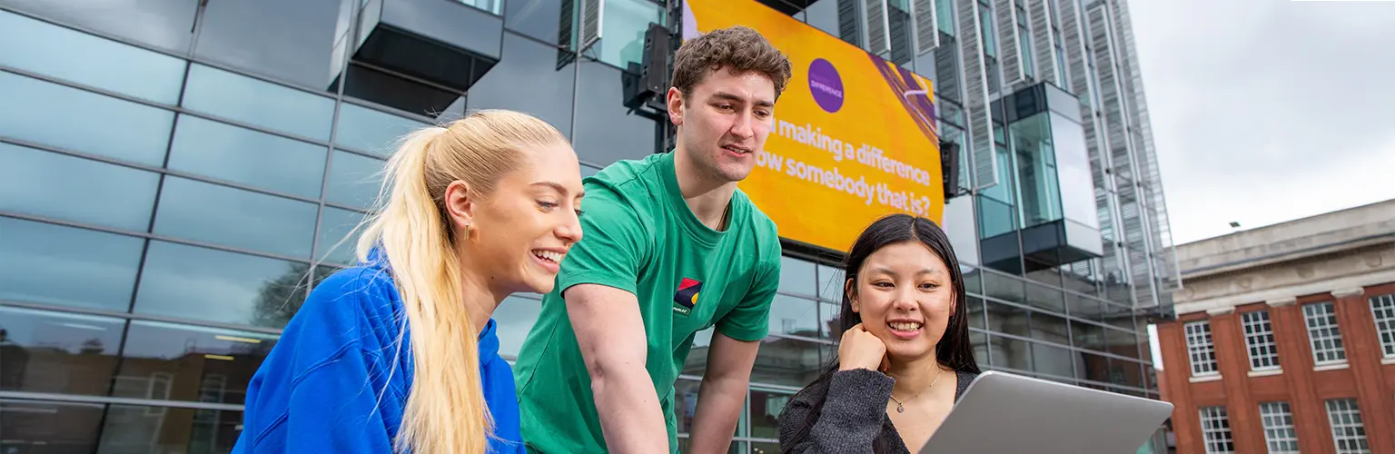 Three students in a university courtyard gathered around a laptop and smiling.