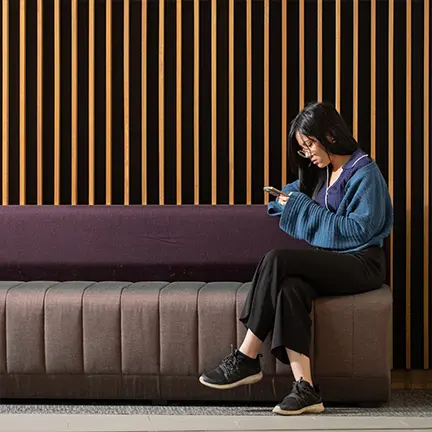 Young woman sitting on a modern bench, focused on her phone, with a wooden slat wall backdrop in a contemporary indoor space.